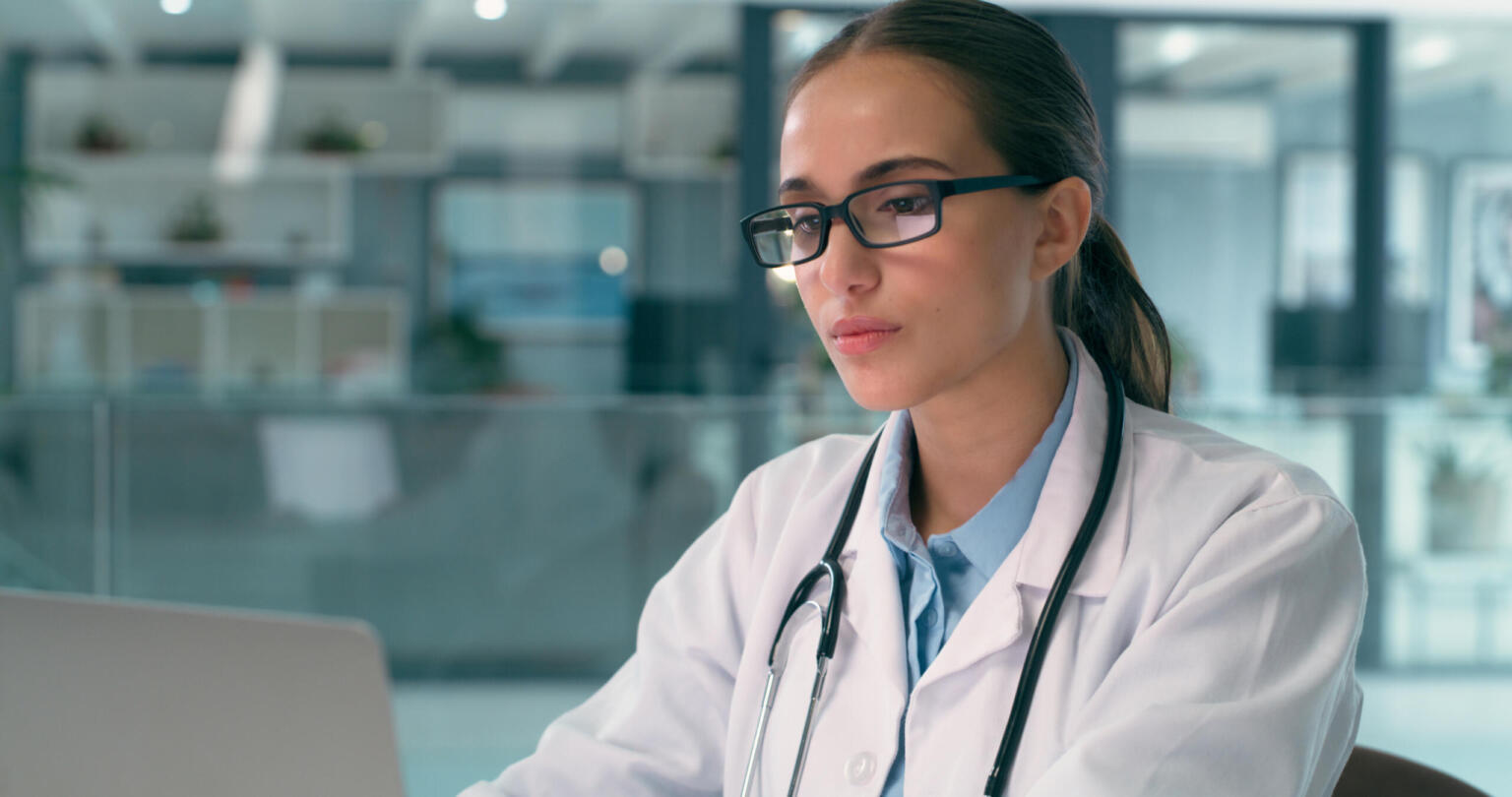 Doctor, woman and laptop for healthcare research, hospital analysis and typing report or review with glasses. medical worker or young person reading clinic news or information on computer in office